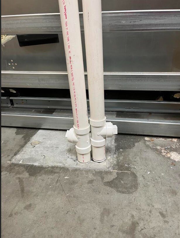 Exposed PVC supply lines emerging from concrete floor, vulnerable to forklift and pallet jack damage — before FST custom fabrication install